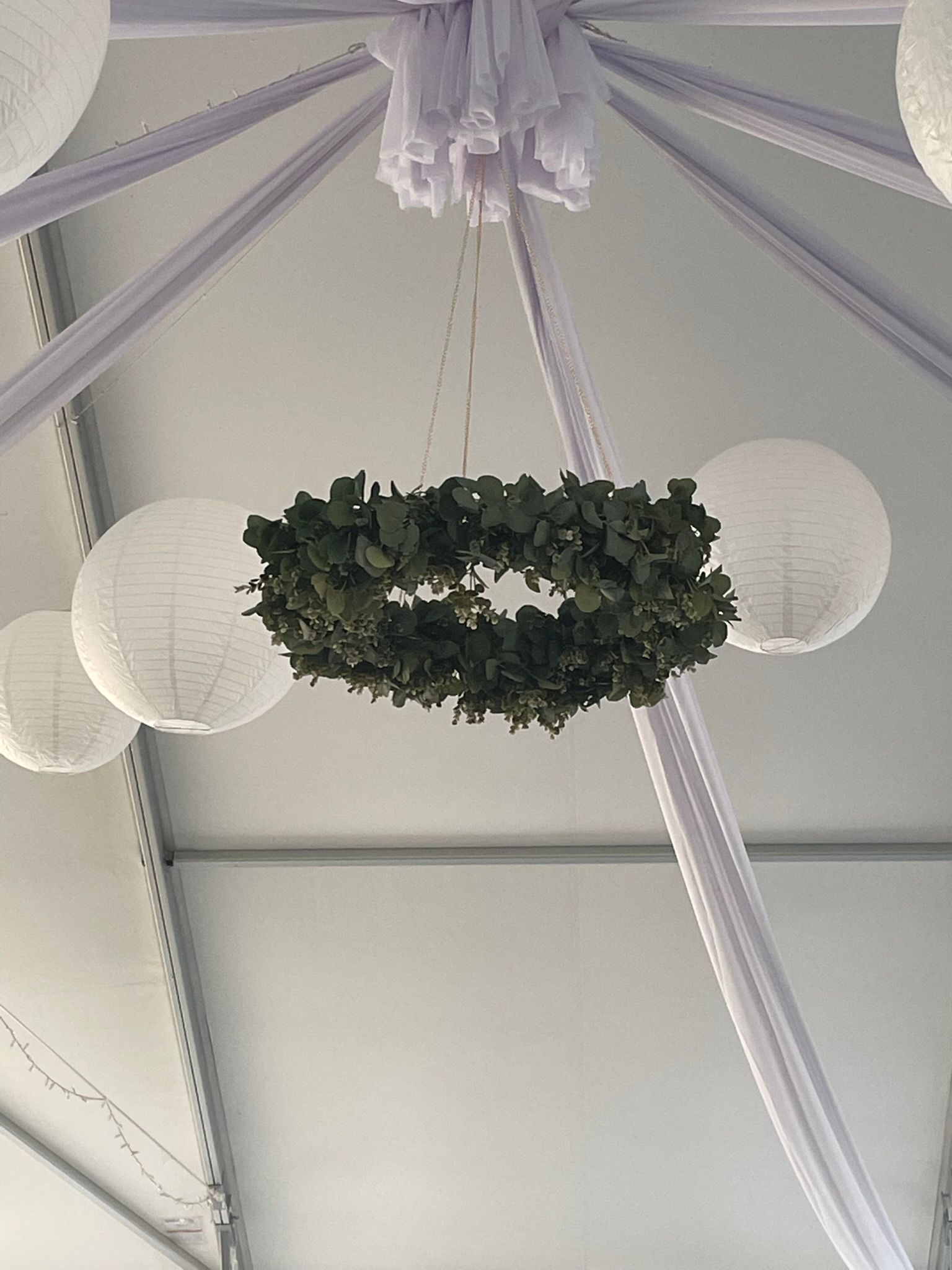 White paper lanterns and a greenery wreath hang from a tent ceiling with lavender fabric accents.