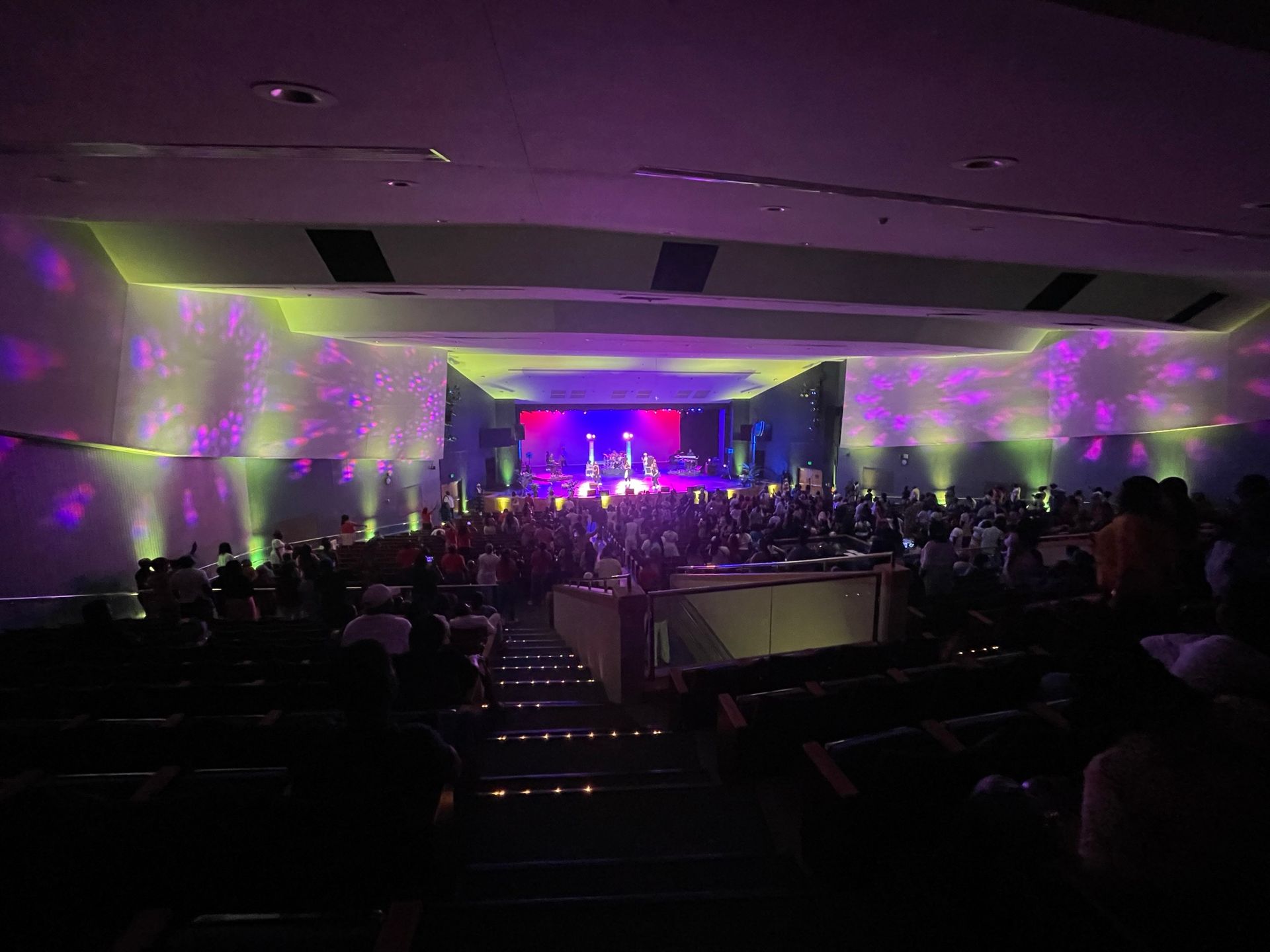 Audience in a dimly lit auditorium watching a performance on a stage lit with purple and green lights.