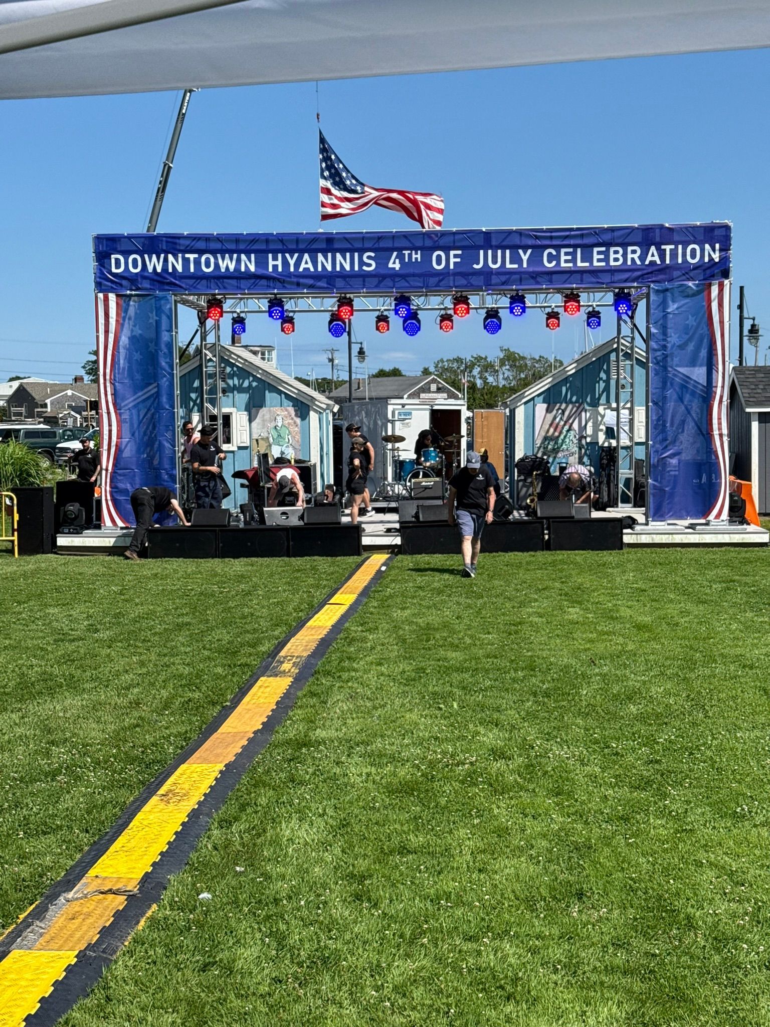Downtown Hyannis 4th of July Celebration with a band on stage, American flag, blue skies, and a grassy area.