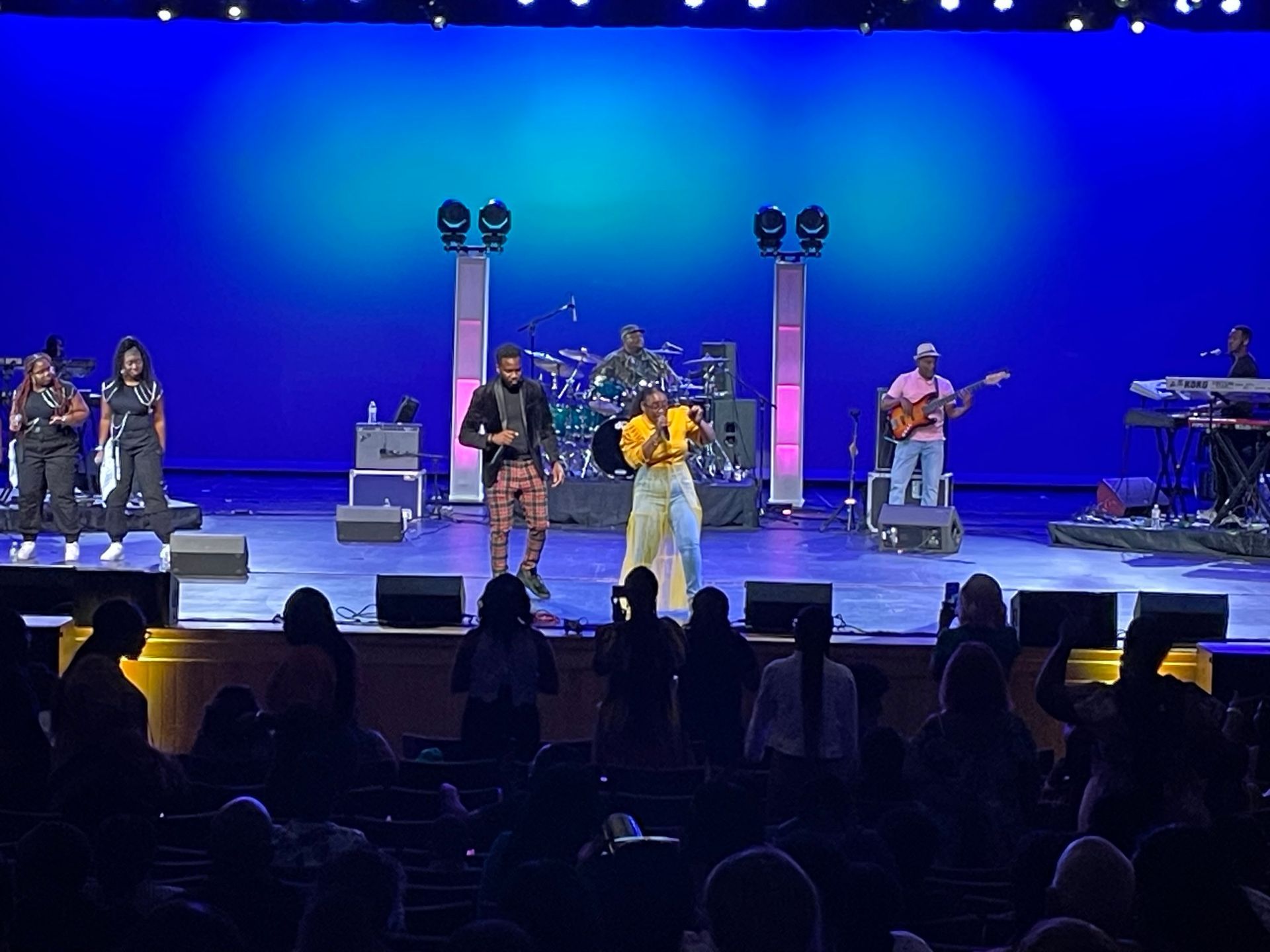 Band performing on stage in a theater, bathed in blue light, with an audience in the foreground.