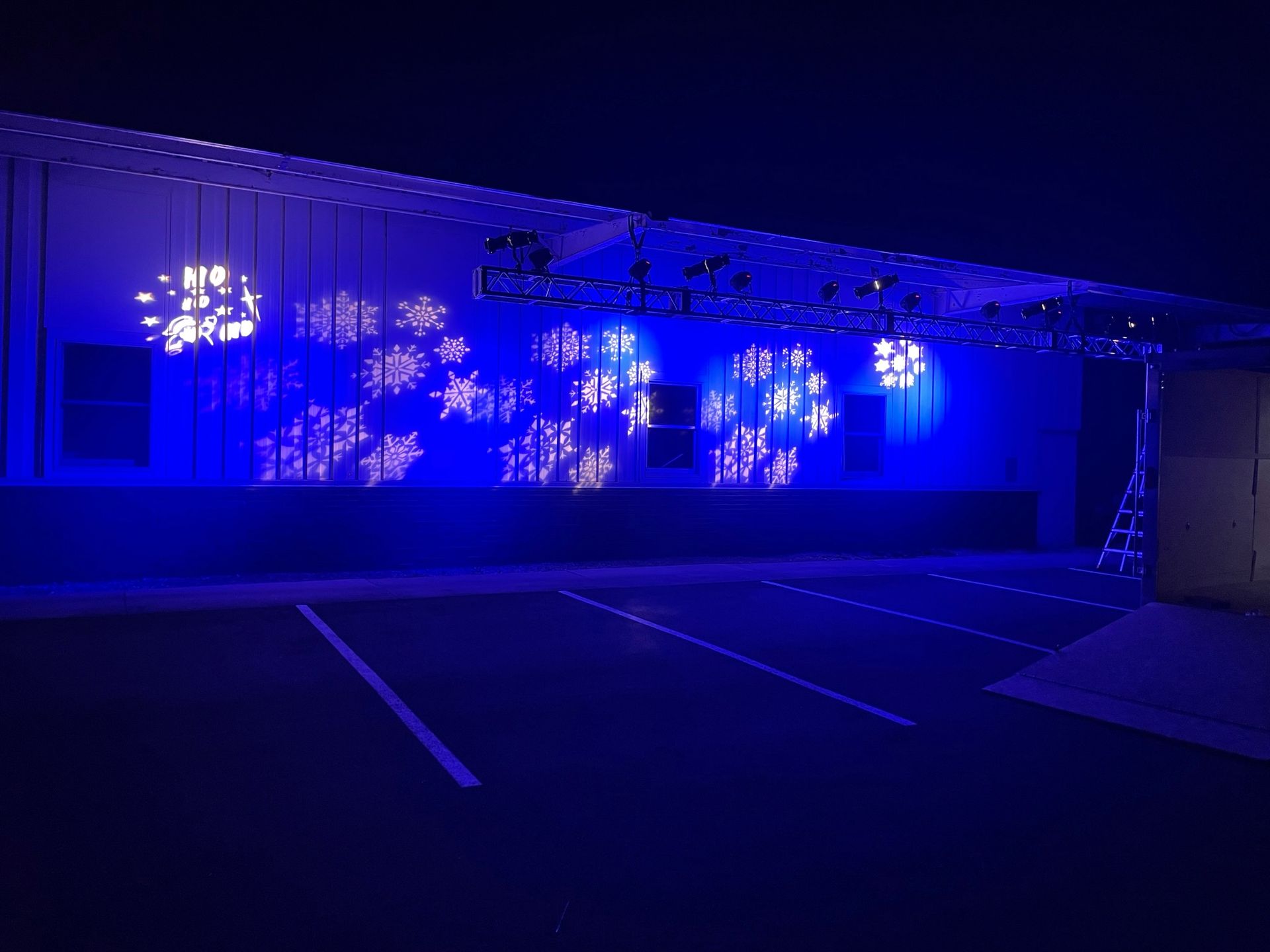 Blue-lit outdoor stage with snowflake light patterns on a white wall. Dark parking lot, metal beams.