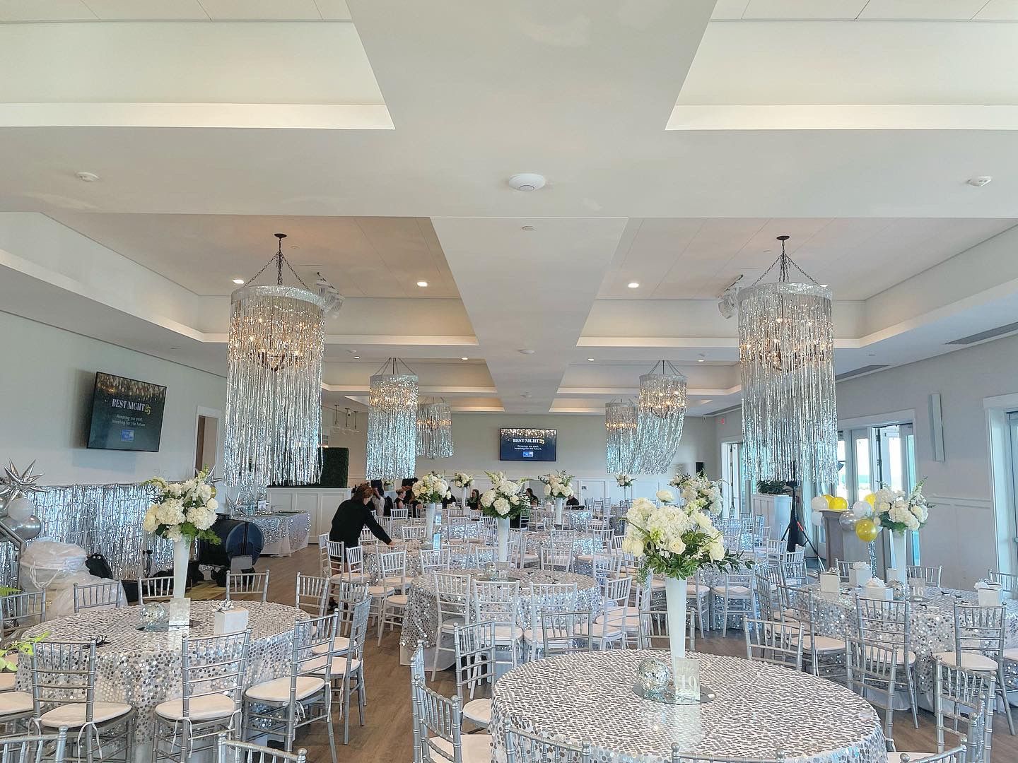 Elegant event hall, silver decor, round tables with white floral centerpieces, chandeliers, empty chairs.