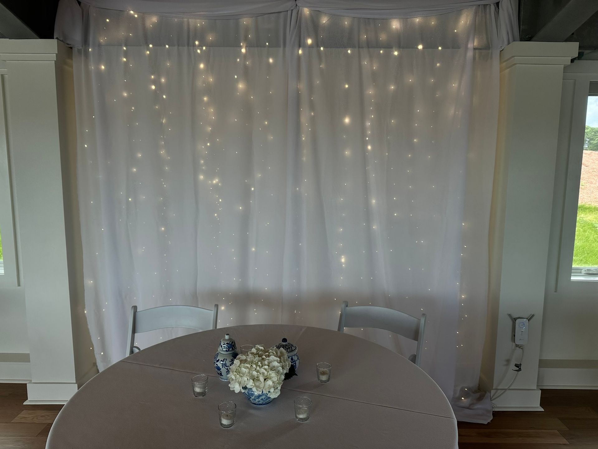 Round table with white chairs, draped with white curtains and fairy lights, with floral centerpiece.