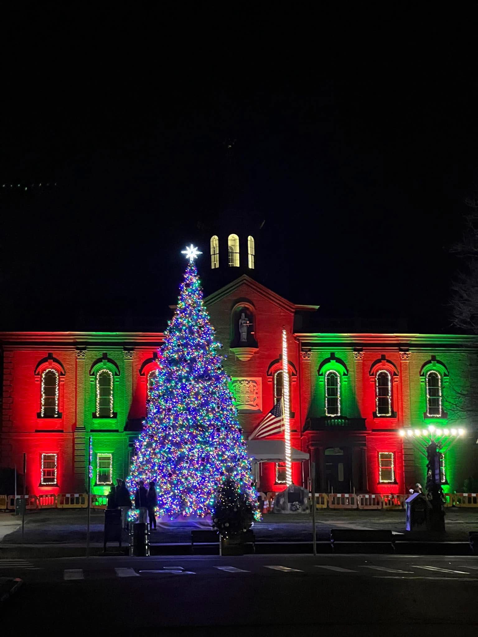 Christmas tree with blue lights in front of a building lit with red and green lights.
