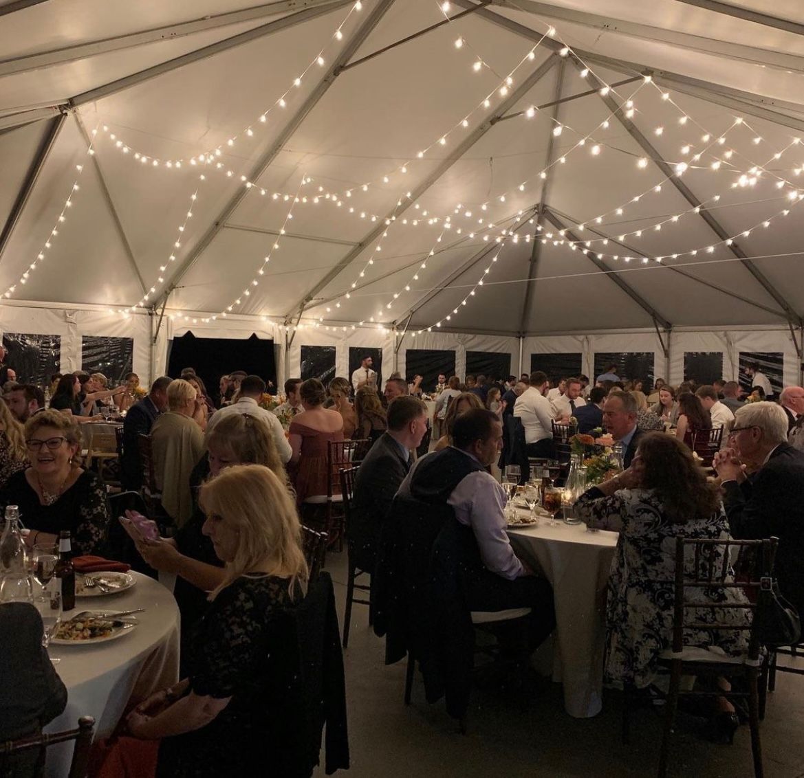 Inside a tent, people dine at tables with overhead string lights.