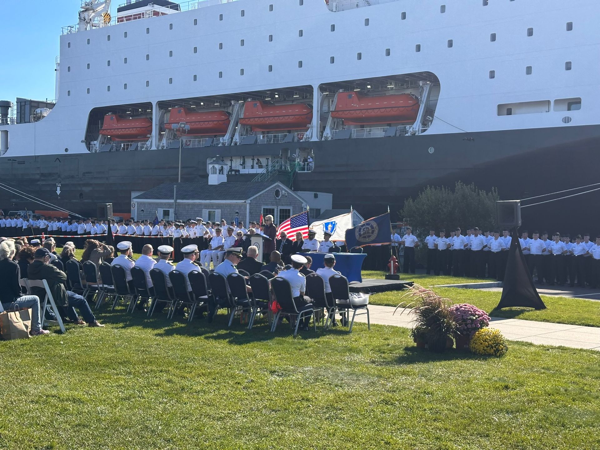 A ceremony on a lawn in front of a large ship. People in uniforms sit and stand.