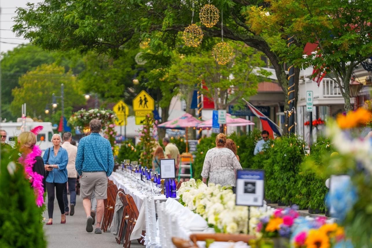 People walking on a decorated street with tables, flowers, and hanging lights.