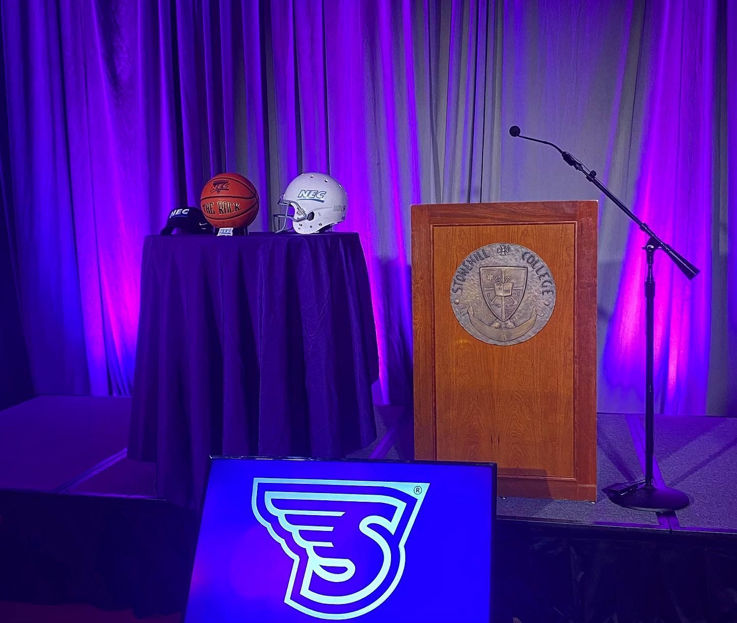 A football and helmet on a table draped in purple, podium with a seal, and a logo in front.