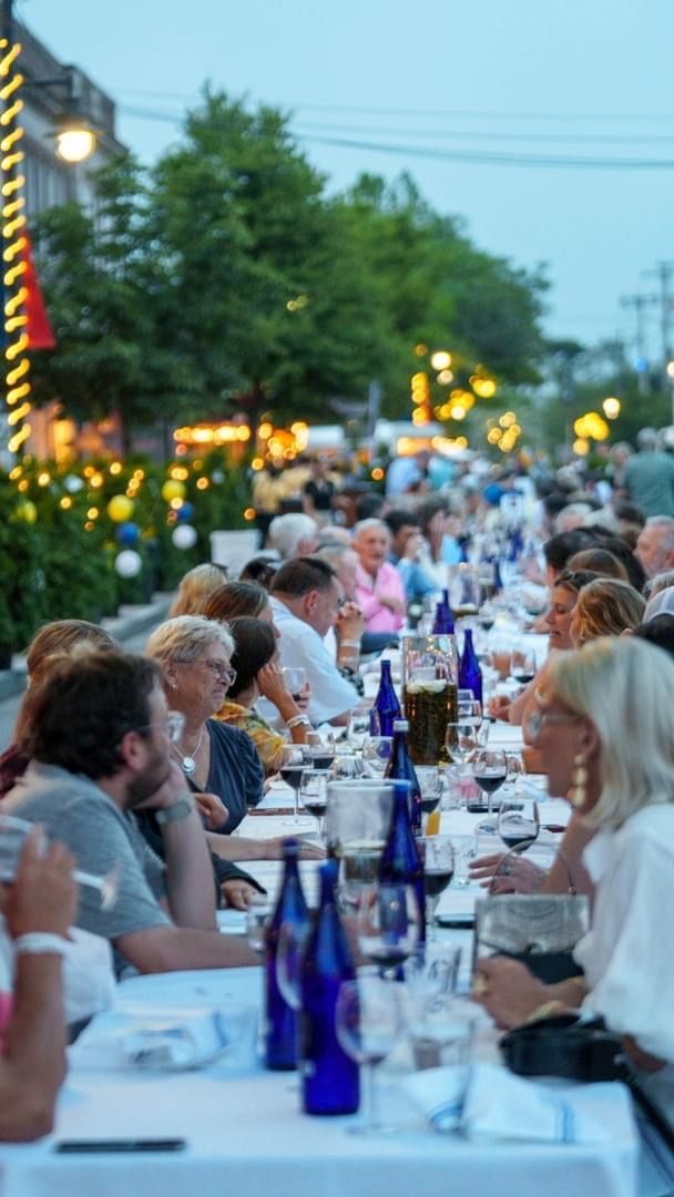 Long table outdoor dinner, people seated, chatting. Blue bottles, white tablecloth. Buildings and trees in background.