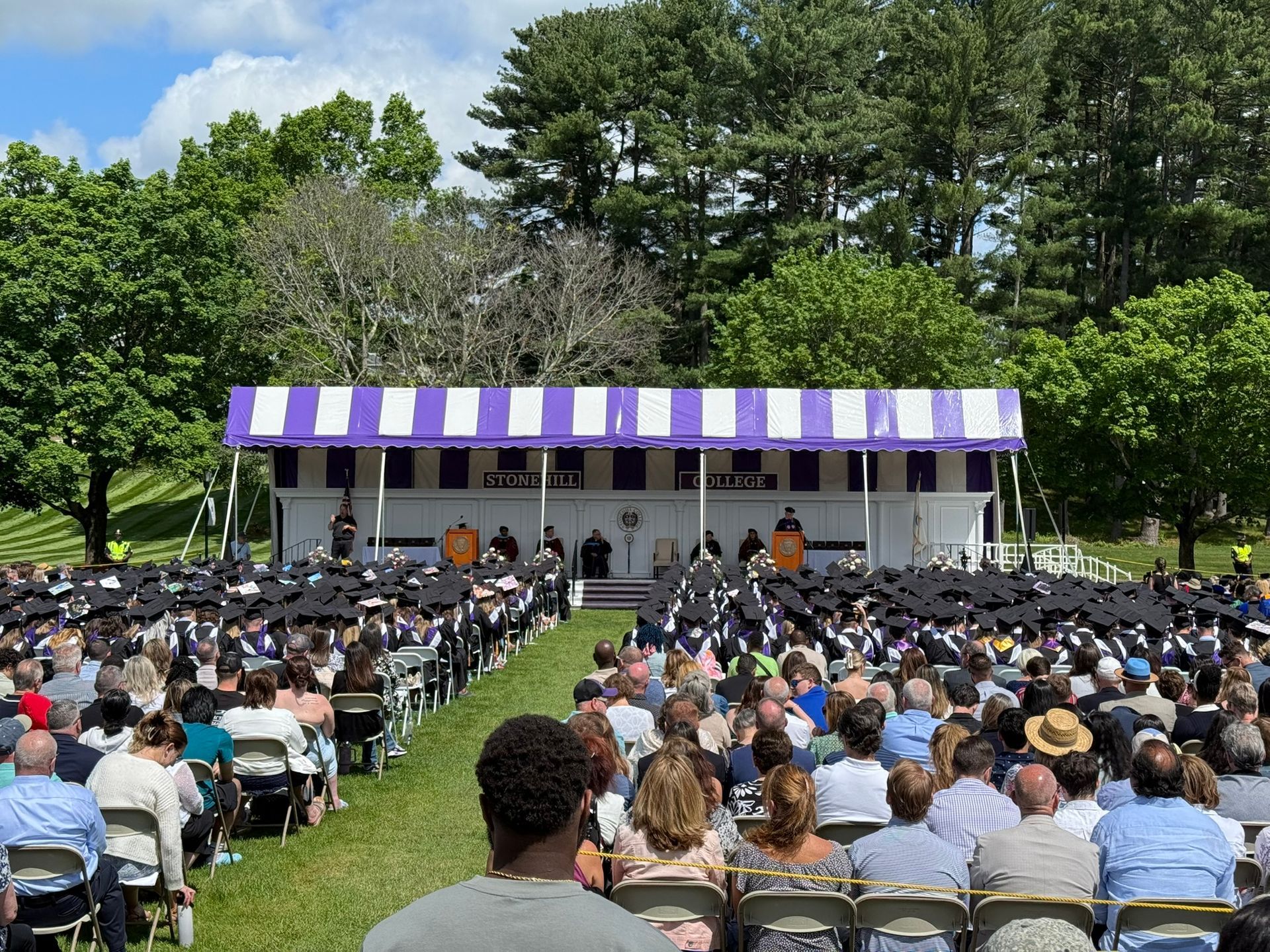 Outdoor graduation ceremony with graduates, audience, and a stage under a purple and white striped tent.