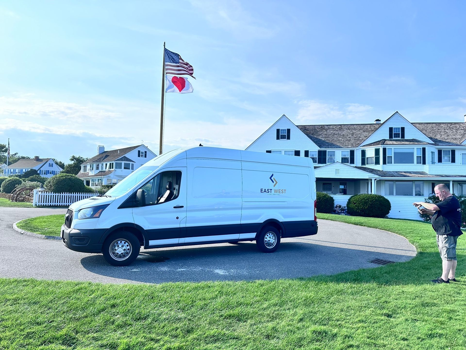 White van parked on a circular driveway, a person taking a picture, a building, and flags.