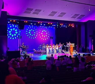 A band performs on stage with purple and blue lighting; audience members in seats.