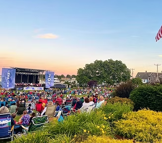 Concert in park at dusk: stage, crowd in chairs, American flag, green grass, yellow flowers, blue sky.