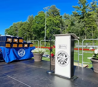Podium on a stage; diplomas on a table with a school logo; outdoor setting with greenery and a track.