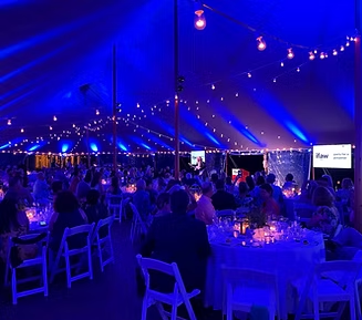 Inside a large tent illuminated with blue lights, tables with seated guests. String lights hang from the ceiling.