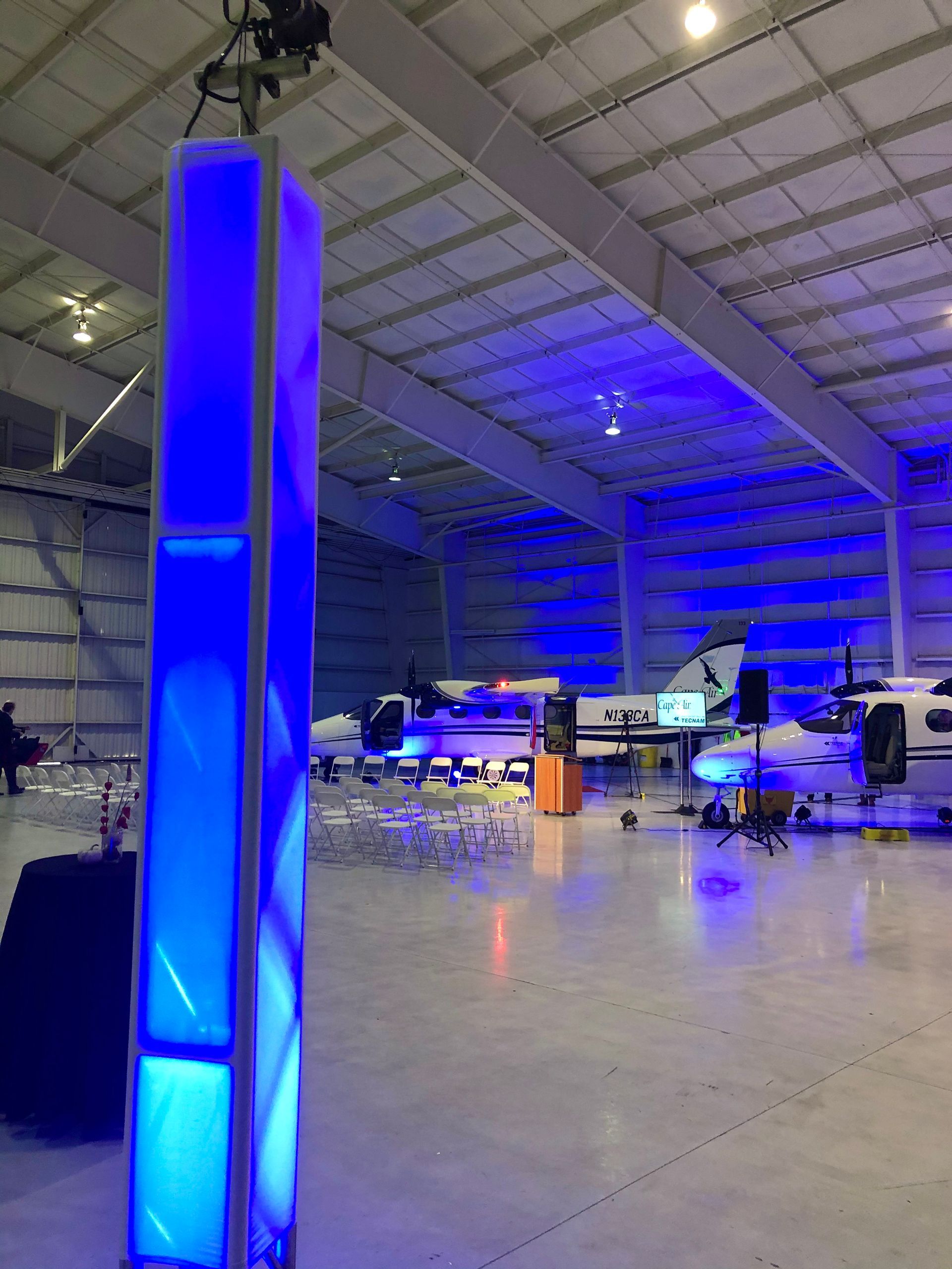 Blue lit pillars in an airplane hangar. Planes and tables are visible.