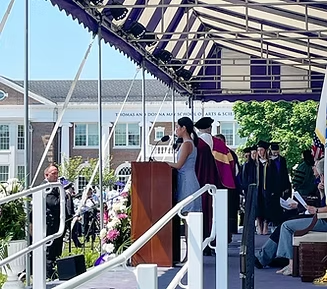 Person speaking at a podium on stage with graduates and a building in the background. Purple and white canopy overhead.