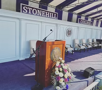 Podium at Stonehill College graduation. Purple carpet, white chairs, and sign.