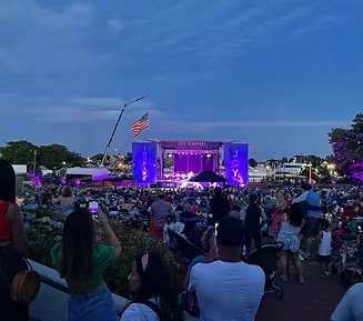 Concert at dusk with a crowd watching a stage lit with purple. American flag visible.