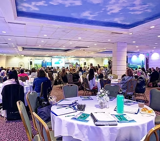 Conference hall with round tables, attendees, and presentations on screens.