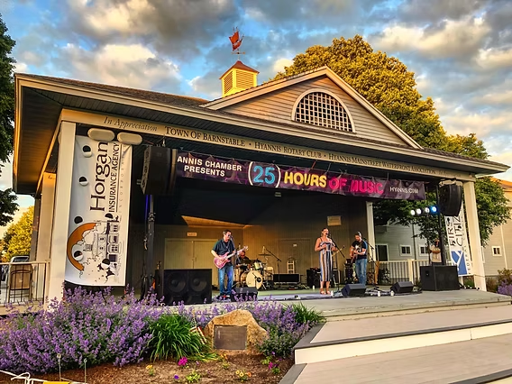 A band performs on an outdoor stage at sunset. Purple flowers frame the stage with a sign that reads 