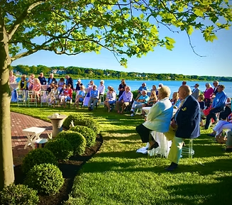 People seated on a grassy lawn facing a body of water during a sunny outdoor event.