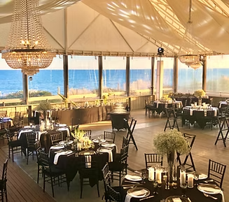Event venue with tables set for a meal, overlooking the ocean. Chandeliers hang from the white ceiling.