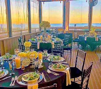 Elegant dining table setting with ocean view at sunset. Black tablecloth, salads, and tall floral centerpieces.