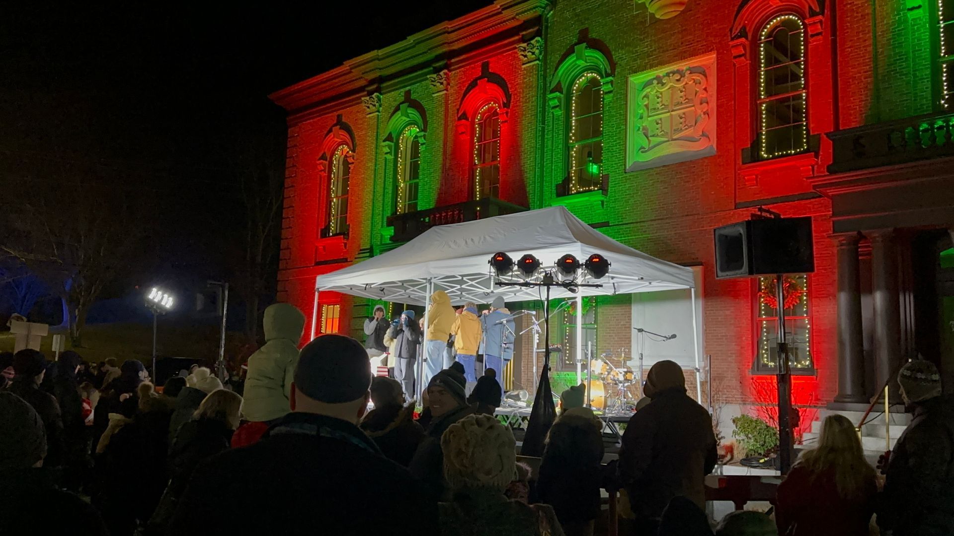 Building lit with red and green lights, stage with performers, and a crowd at night.