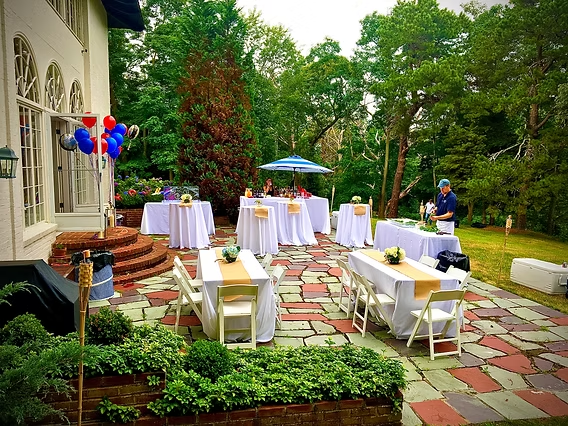 Patio party with tables, white linens, and guests. Green trees and a house in the background.