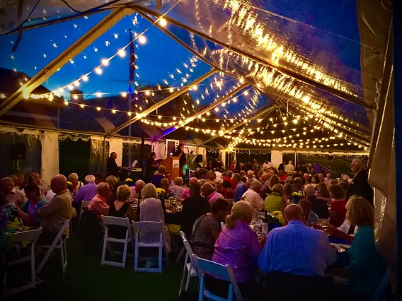 Outdoor event under a clear tent with string lights. A crowd of people sit at tables.