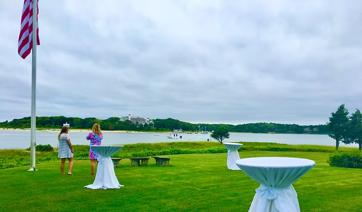Lawn party overlooking water; American flag, two people near white cocktail tables, cloudy sky.