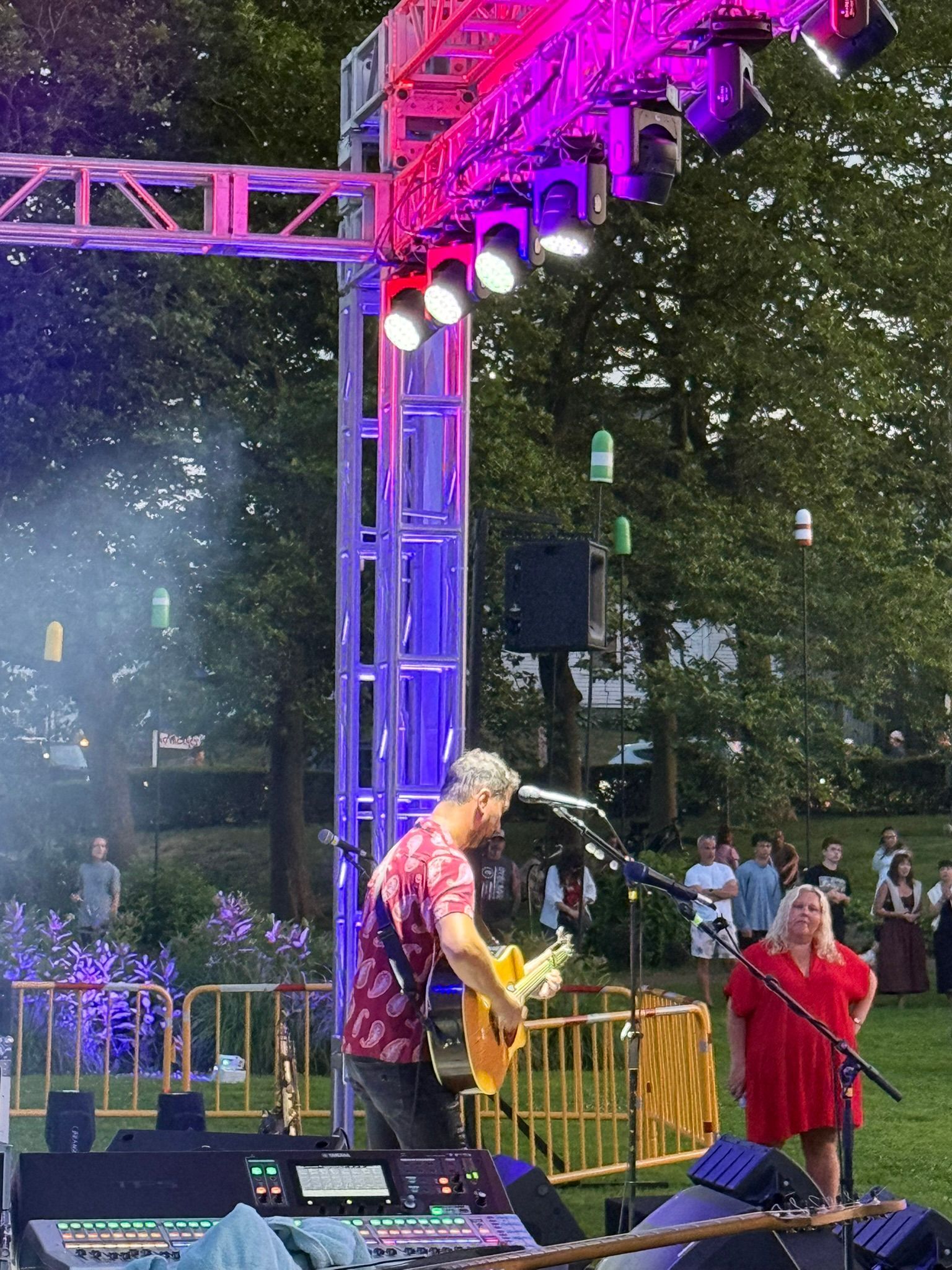 Man playing guitar on stage with lighting rig. Woman in red dress watches. Outdoor concert.
