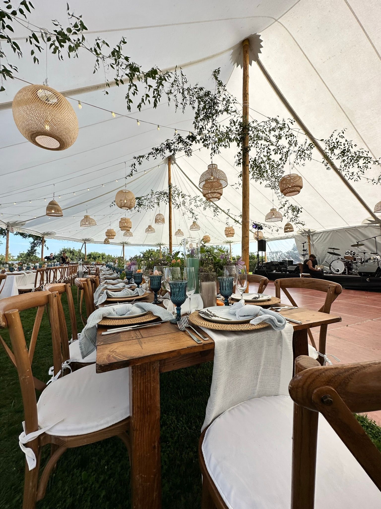 Long wooden tables set for a celebration inside a white tent, decorated with hanging woven lanterns and greenery.