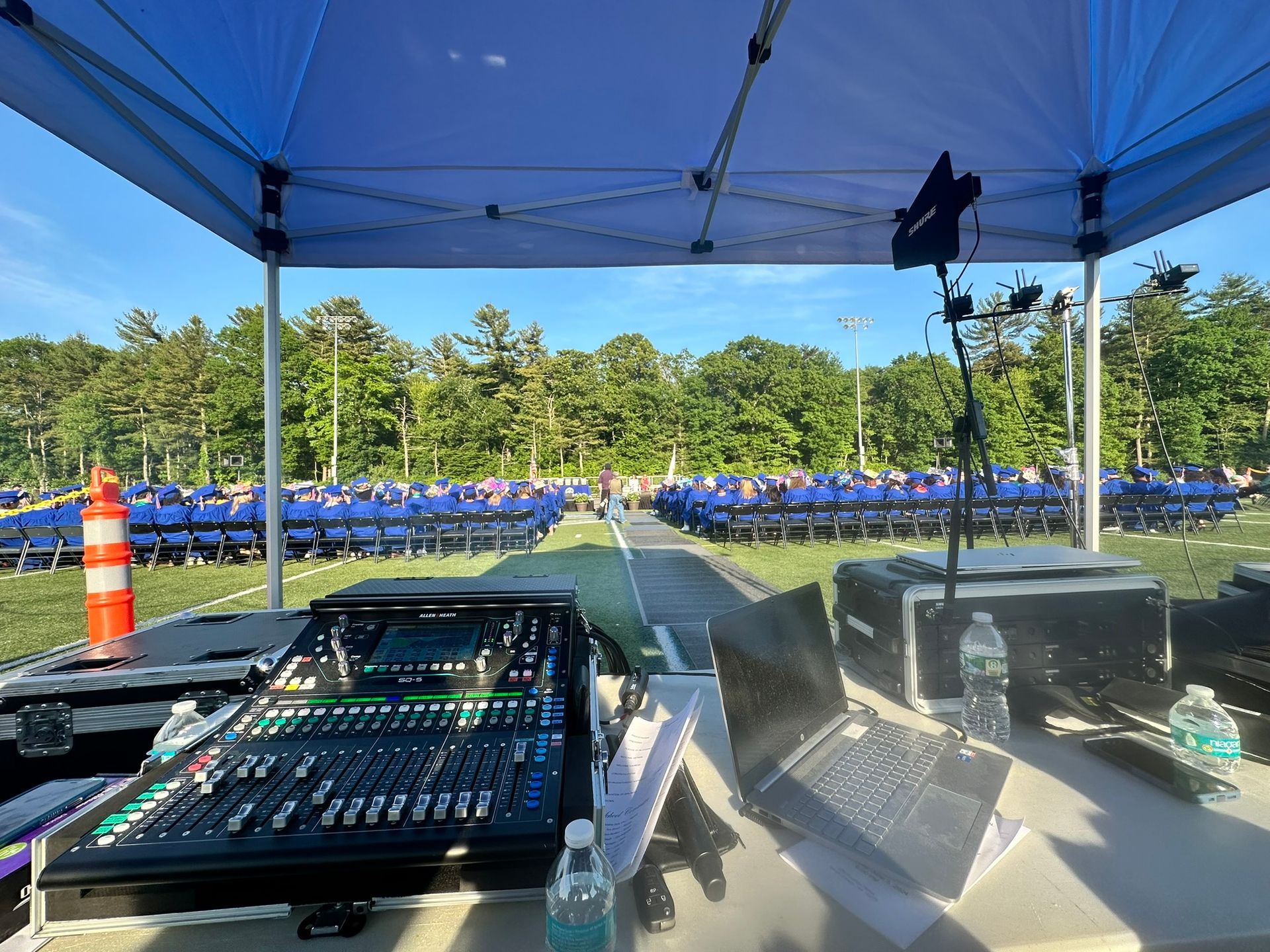 Sound equipment setup at outdoor event. Rows of blue chairs arranged, forest background under a blue sky.