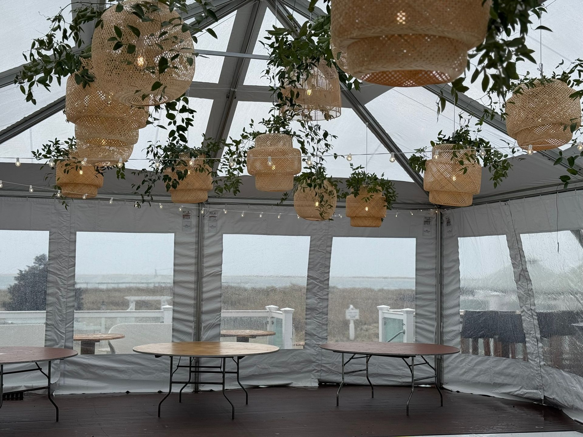 Clear tent interior with hanging woven lights and greenery, tables set, ocean view through windows.