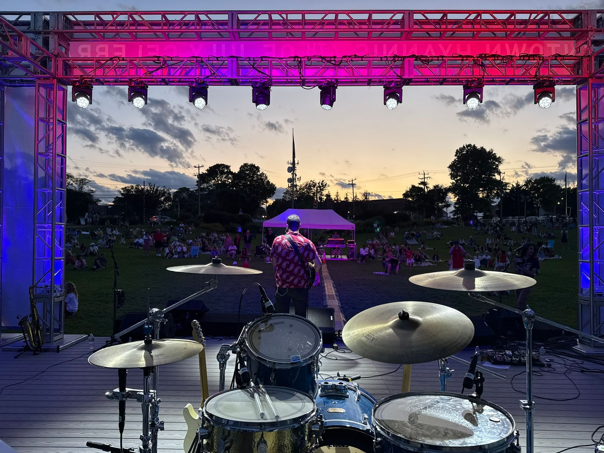 Drum set on stage at an outdoor concert, lit by pink and blue lights, with a guitarist playing.