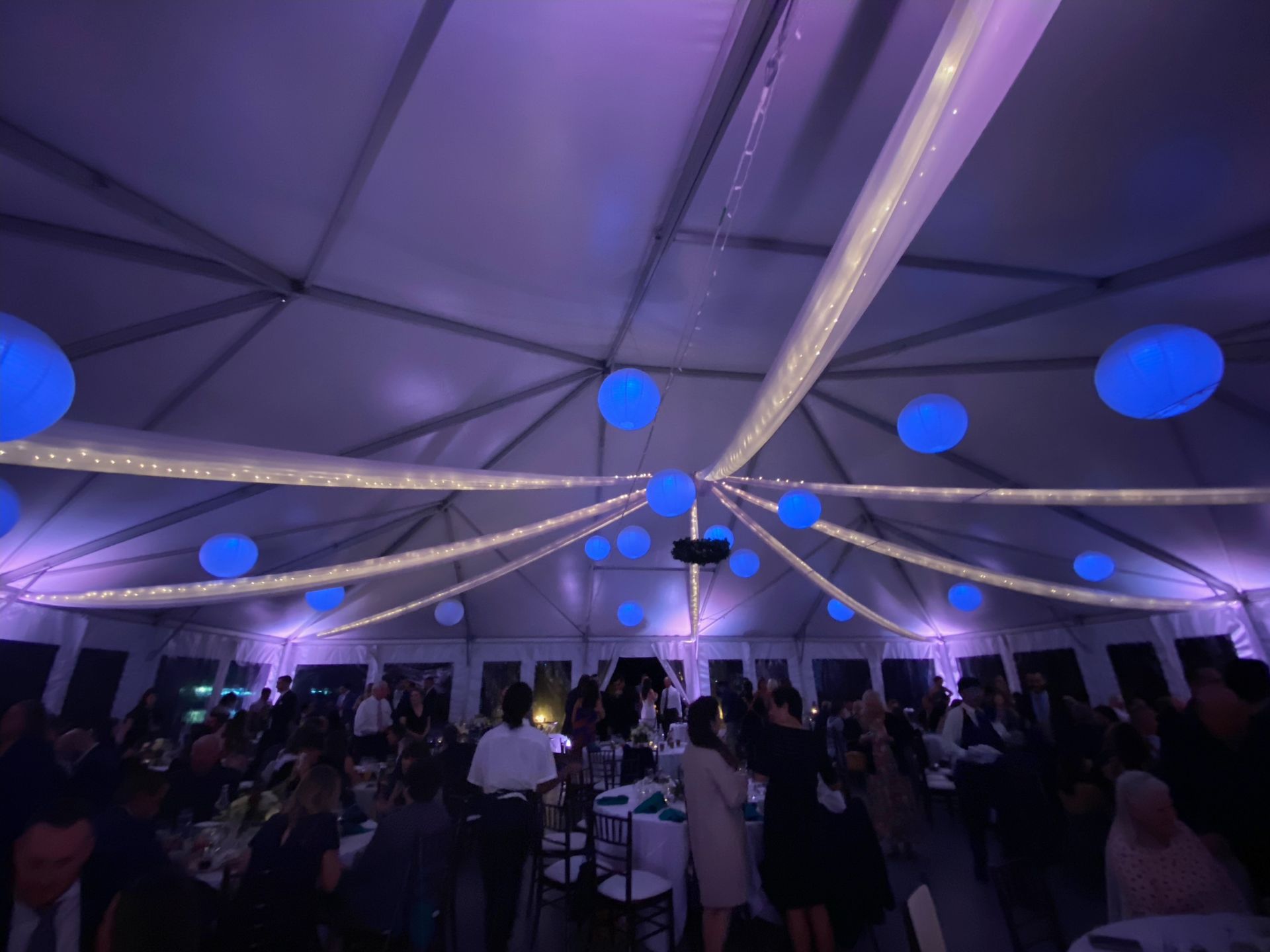 Event tent with blue lighting, string lights, and floating spheres. People are gathered, possibly at a party or reception.