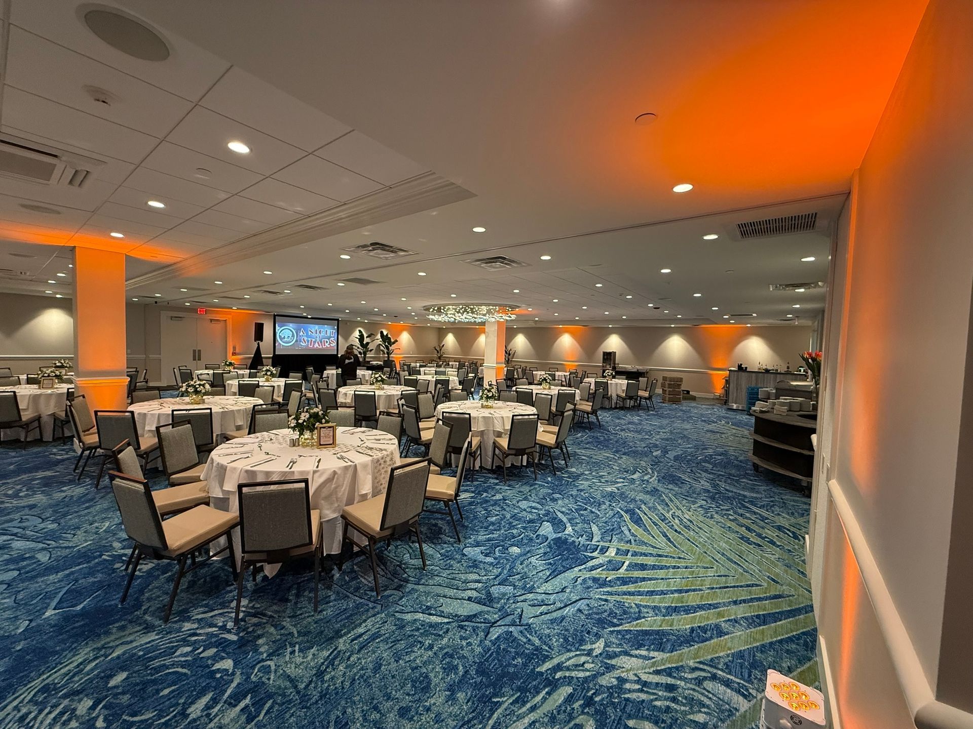 Banquet hall with round tables set for a gathering, blue patterned carpet, orange uplighting.