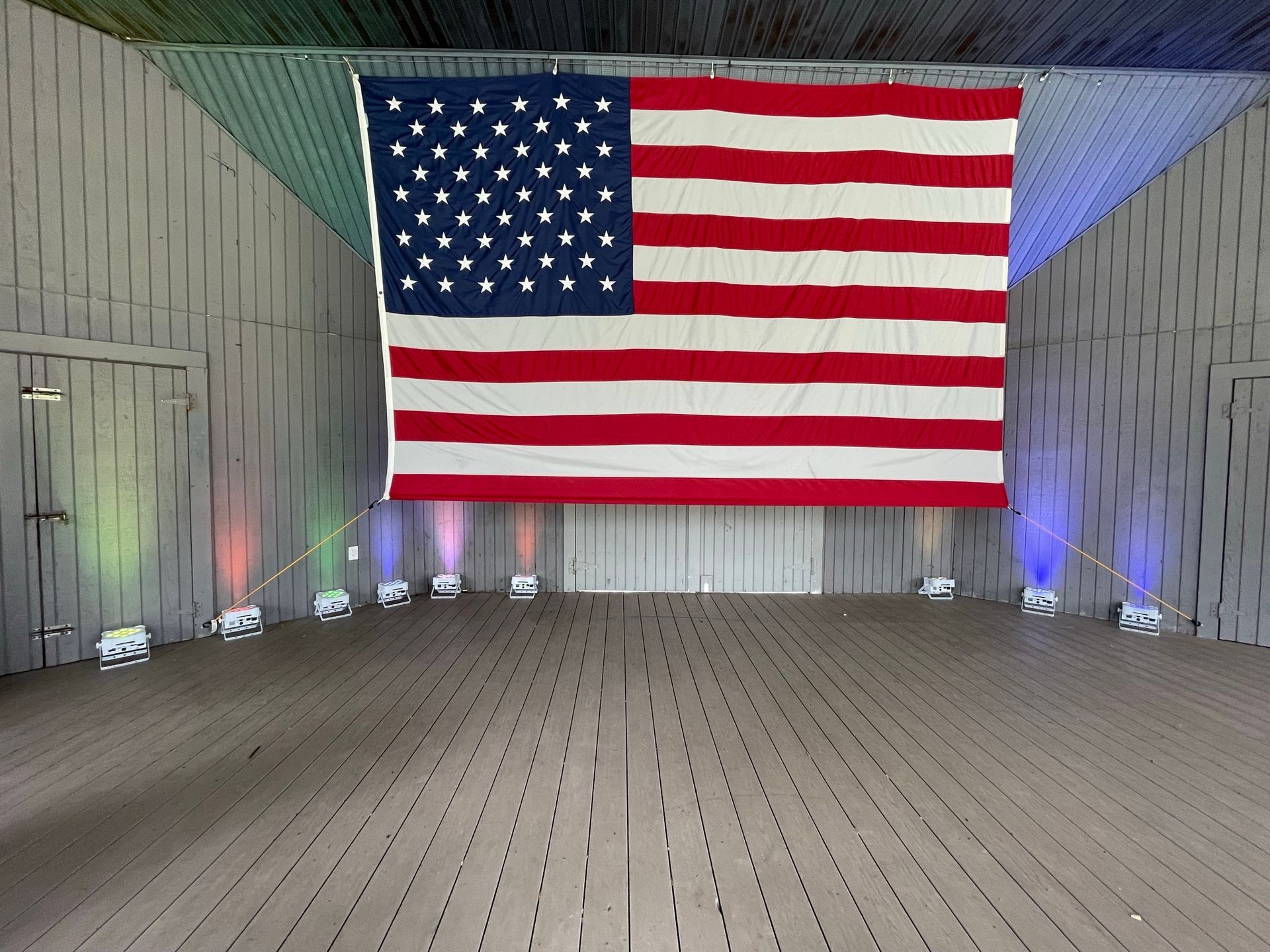 Large American flag hanging inside a barn-like structure, with stage lights on the floor.