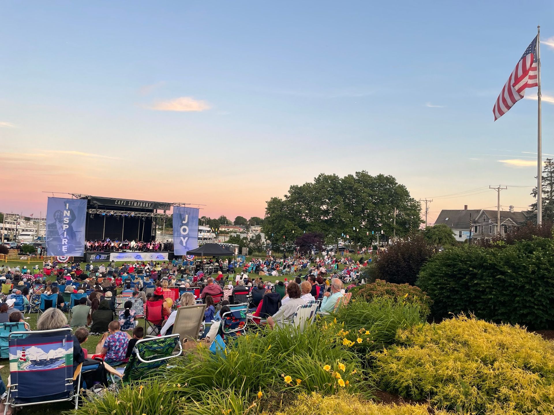 A concert on a grassy field at dusk with a stage, audience, and American flag.