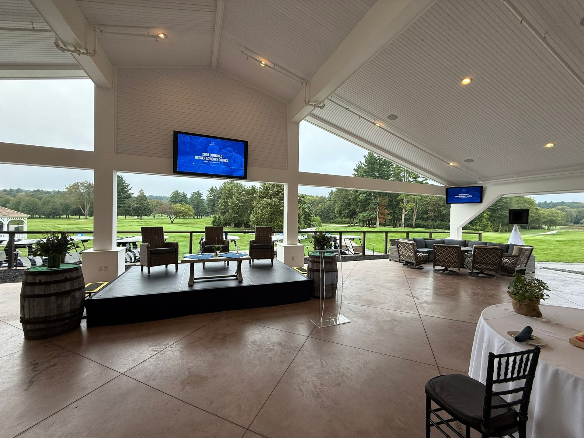 Covered patio with a stage, chairs, and two TVs overlooking a golf course.