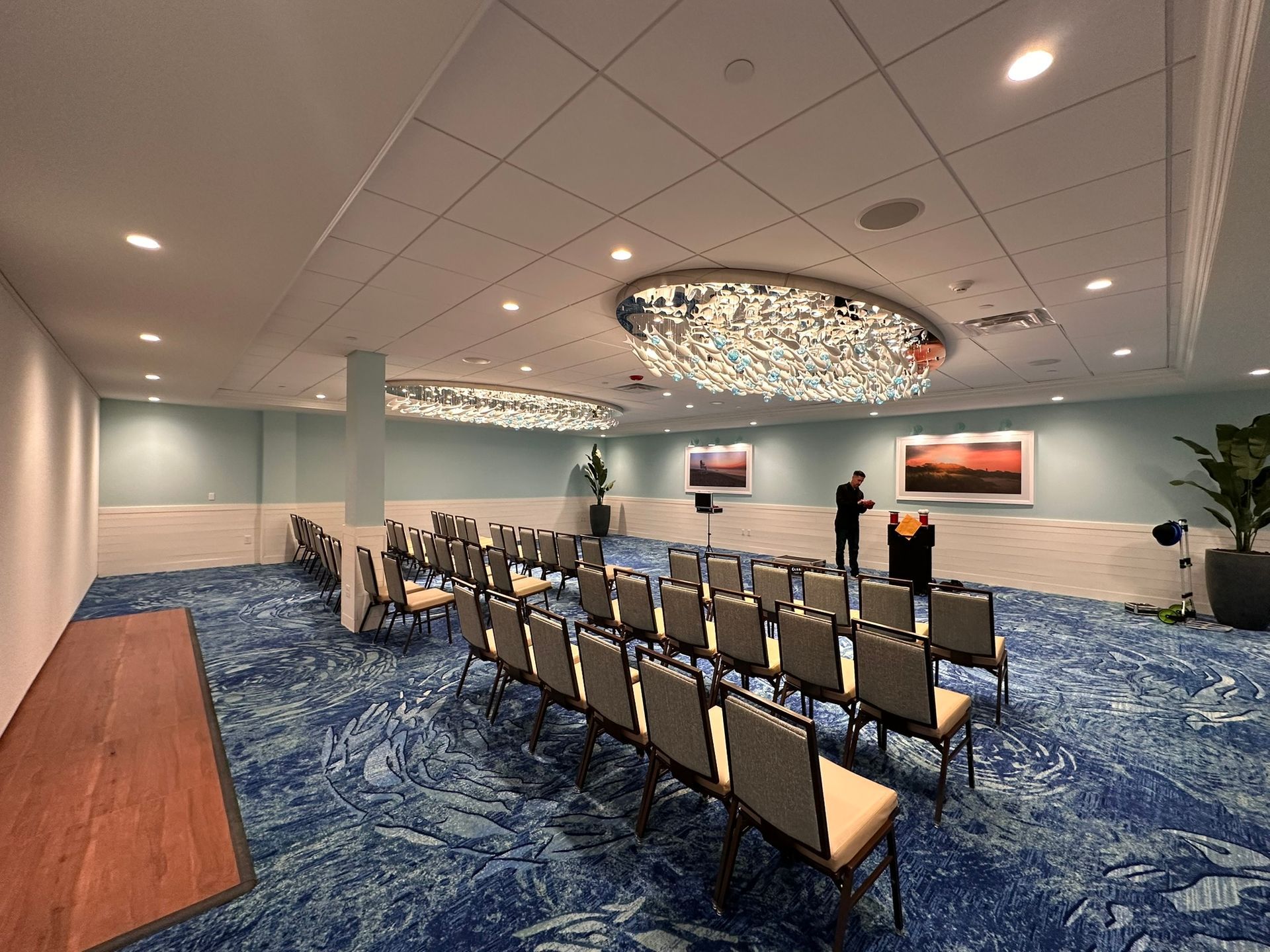 Auditorium with rows of chairs, presentation setup. Two people stand near a podium, art on walls, blue carpet.