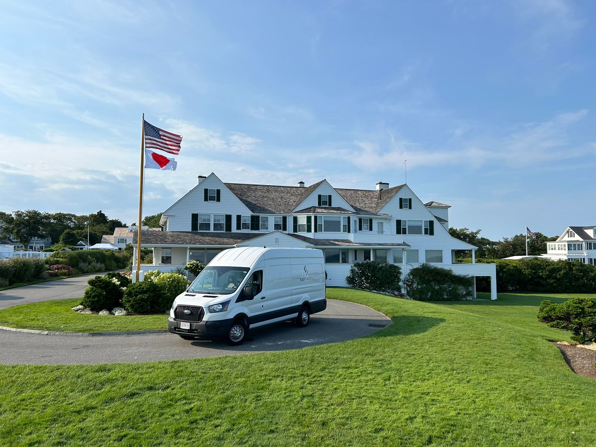 White van parked on driveway of a large white house with an American flag. Blue sky.