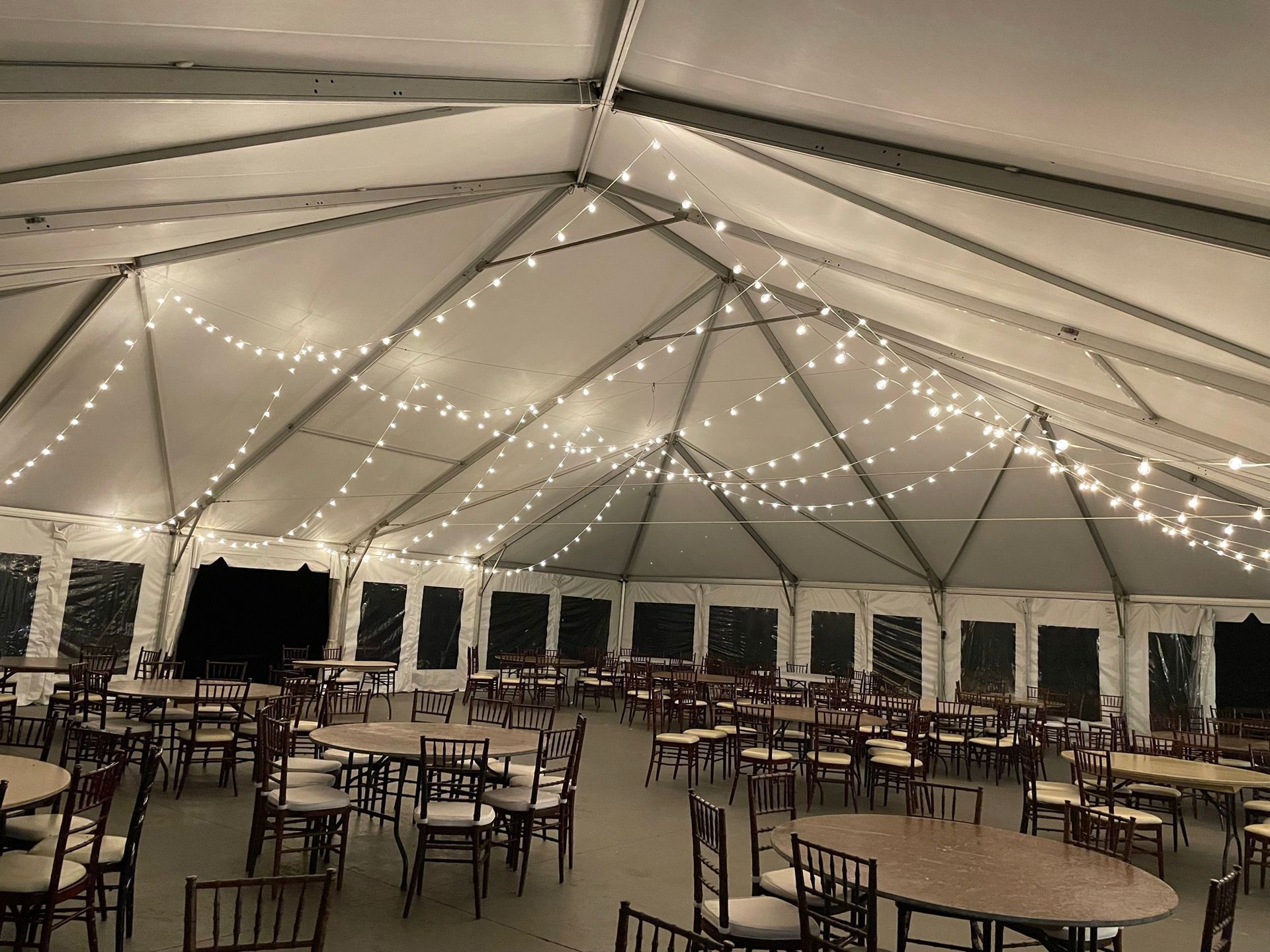 Inside of a large tent, set up with round tables, chairs, and string lights.