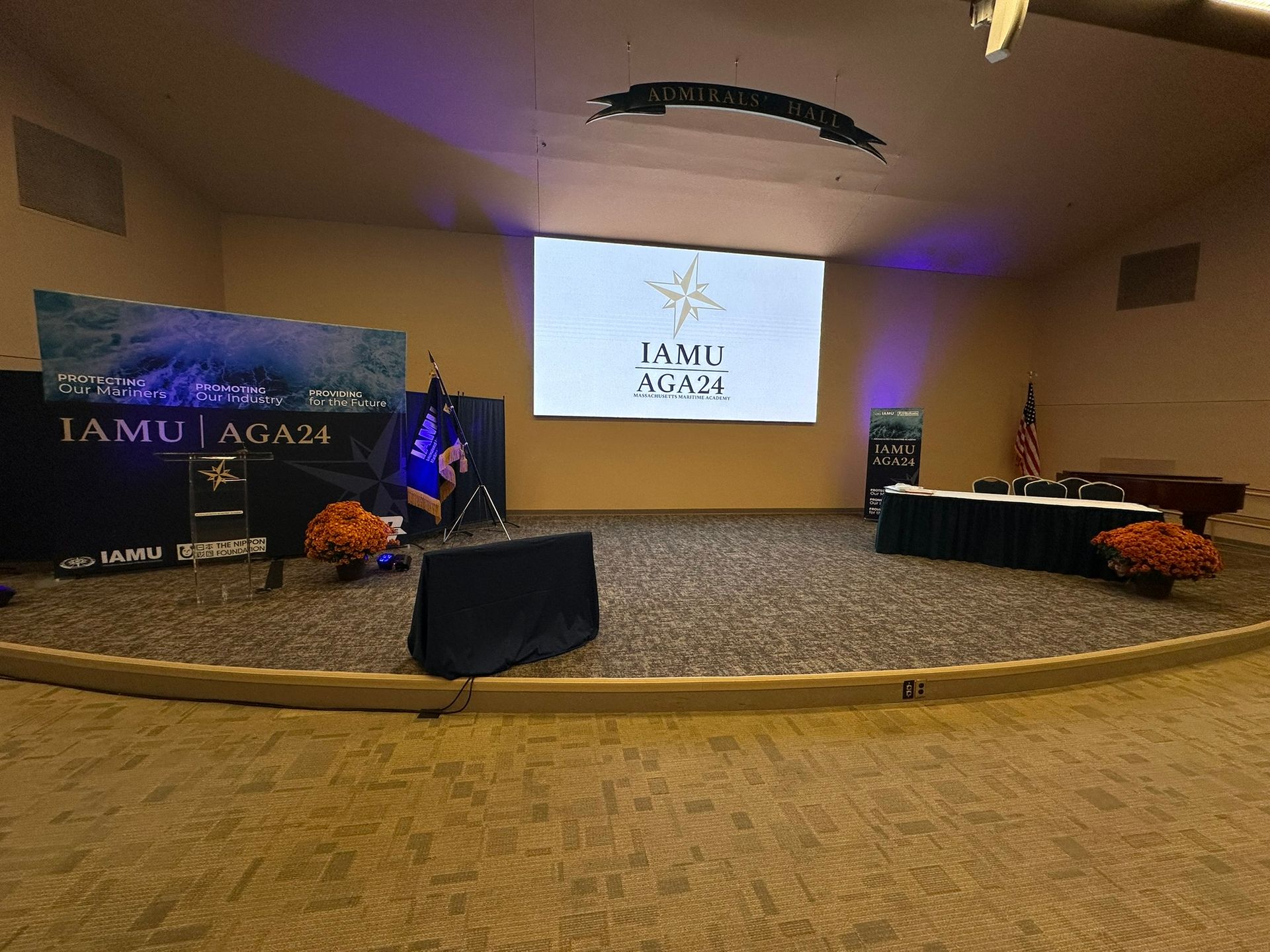 Stage set for an event;  a screen displays a logo. Tables with decor and US flag on the right.  A podium and backdrop on the left.