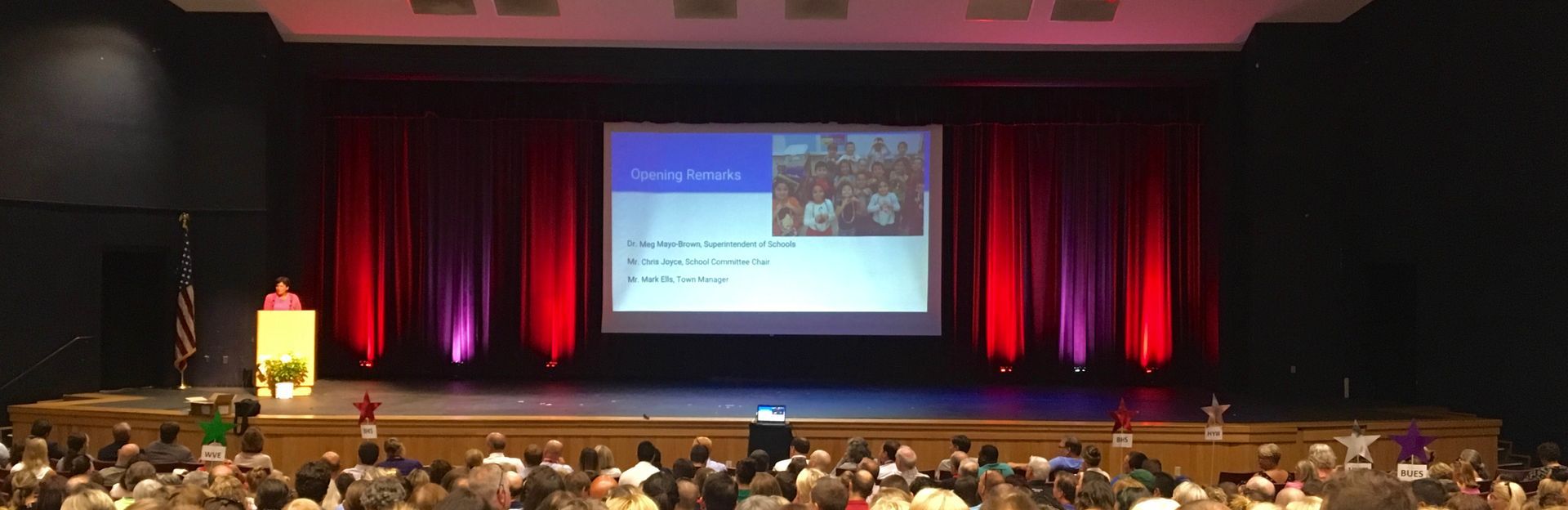 A stage with a screen displaying a presentation, a speaker at a podium, and an audience in a large hall.