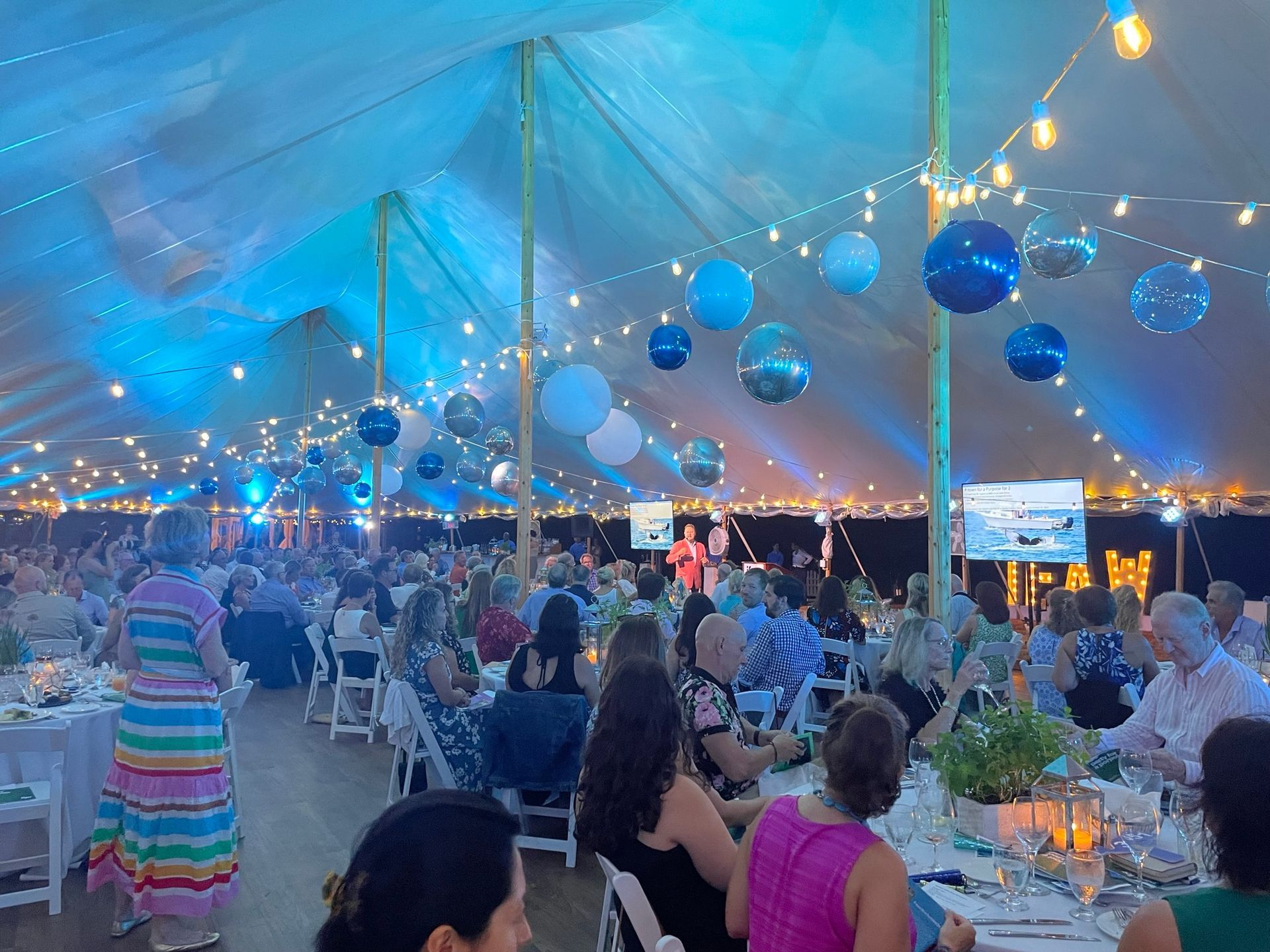 Event in a tent with guests seated at tables, decorated with blue lights and balloons.