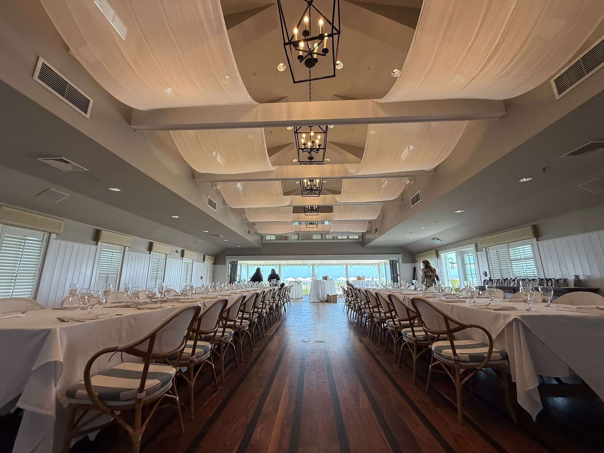 Long dining room with tables set for a meal, draped ceiling, rattan chairs, and large windows.
