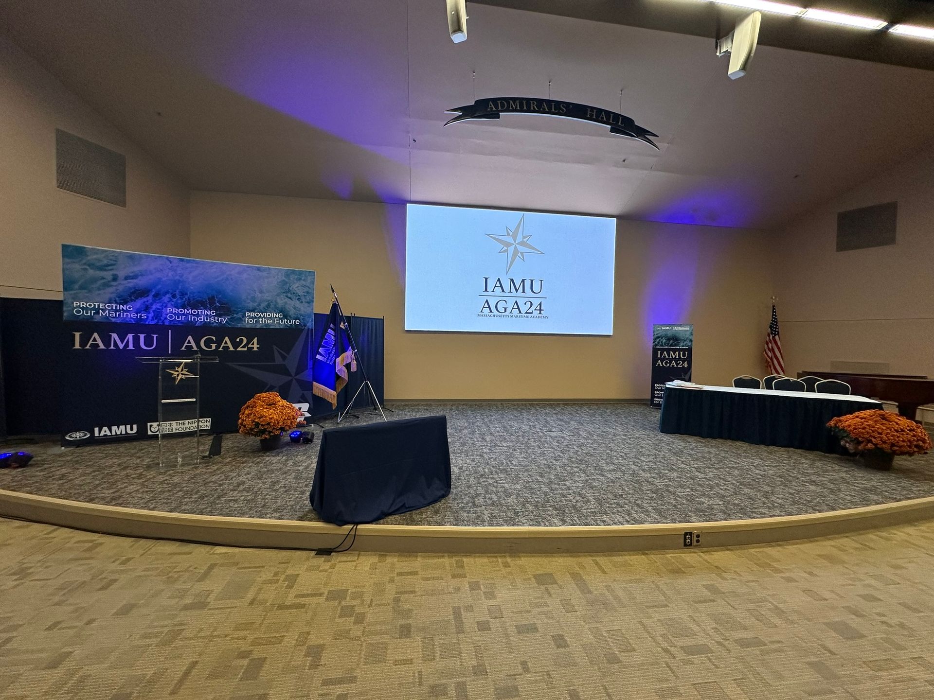 Stage setup for an event: stage, screen with logo, draped tables, US flag, and floral arrangements.